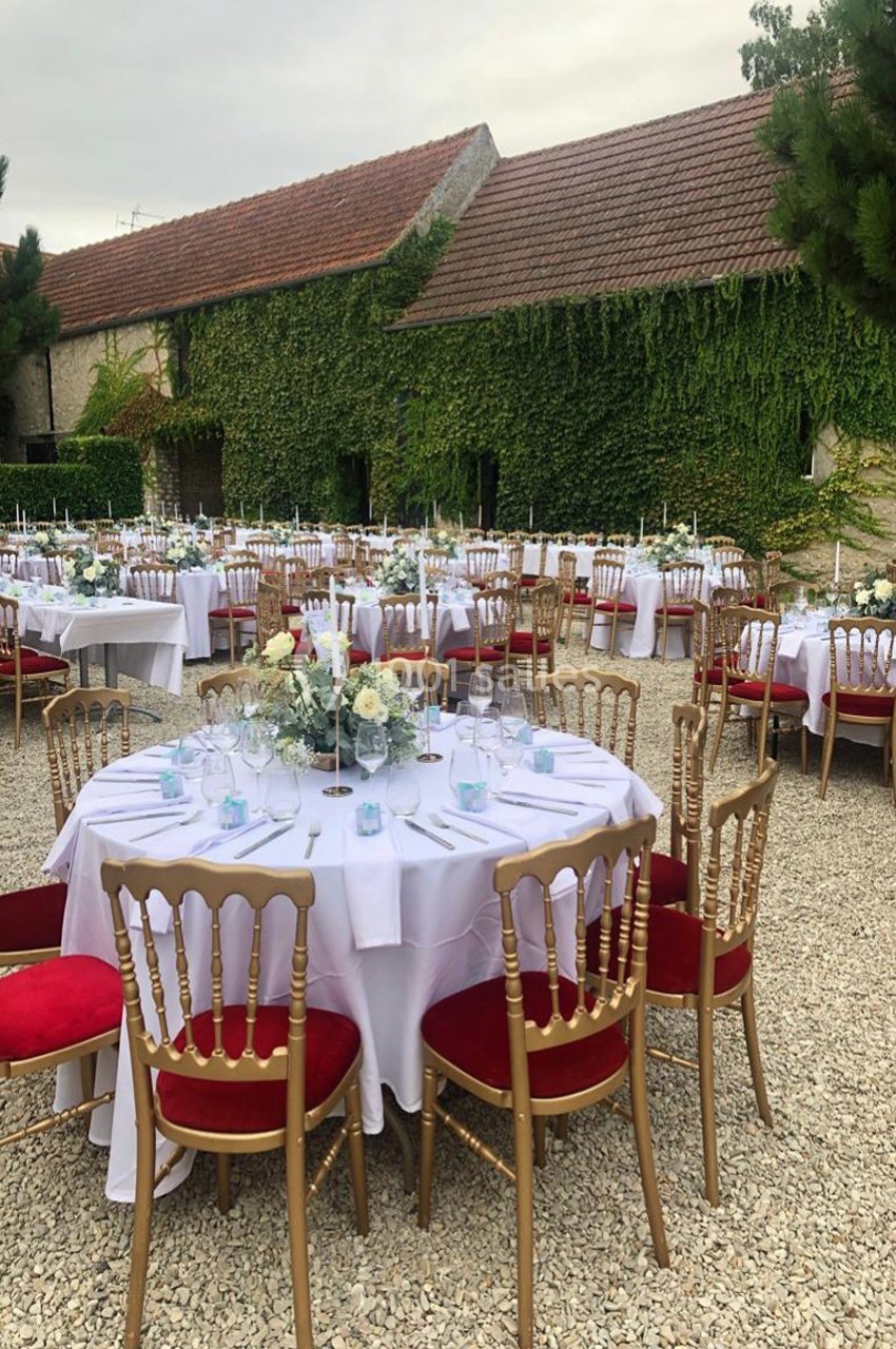 Tables rondes dressées avec nappes blanches, chaises dorées à coussins rouges, disposées en extérieur près d'un bâtiment…