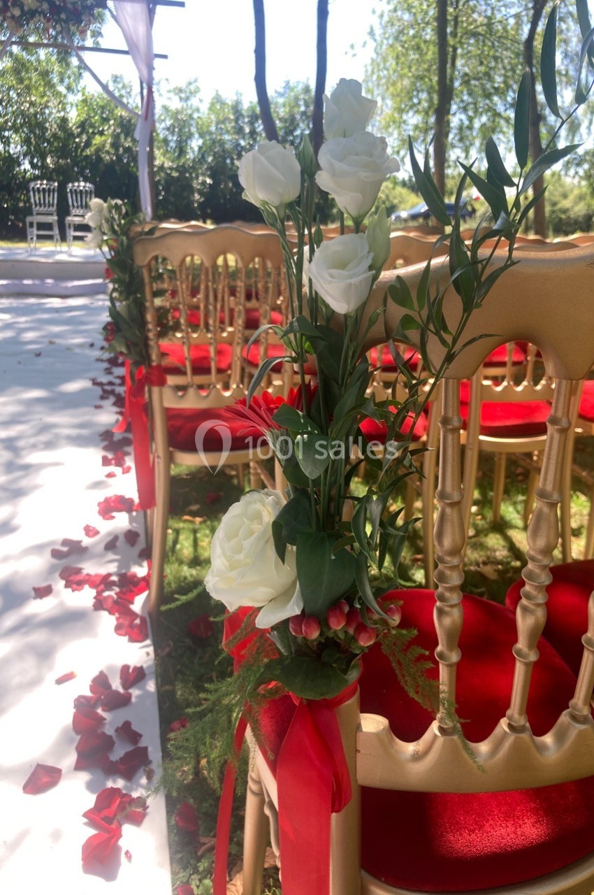 Chaises dorées décorées de fleurs blanches et rubans rouges alignées pour une cérémonie en extérieur.