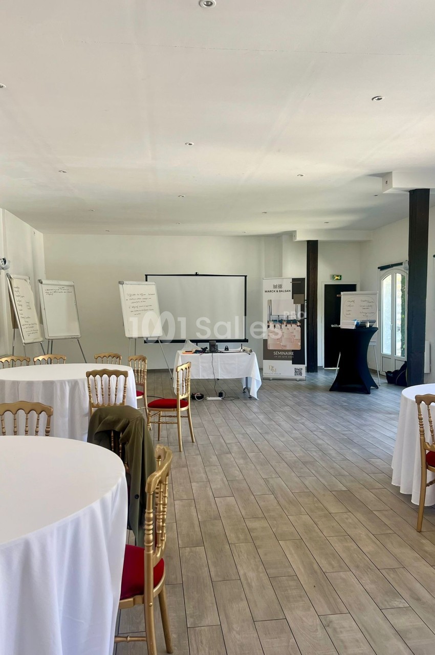 Salle de réunion lumineuse avec tables rondes, chaises, paperboards, écran de projection et grandes fenêtres.