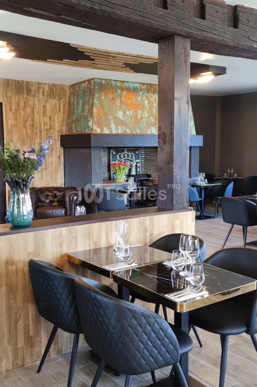 Salle de restaurant moderne avec tables dressées, chaises noires matelassées et cheminée en arrière-plan.