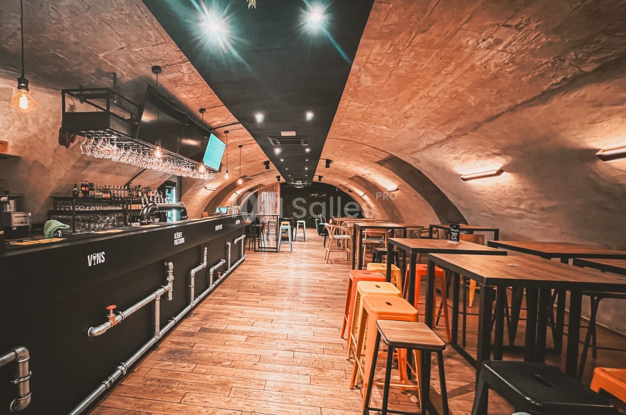 Intérieur d'un bar avec un long comptoir, des tables hautes et des tabourets, dans une salle voûtée en pierre.