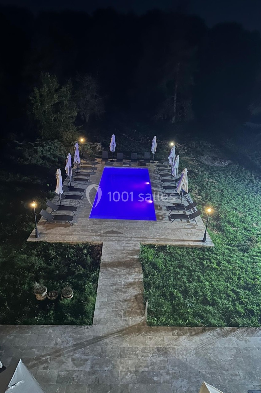Piscine éclairée en bleu entourée de transats et parasols fermés, située dans un jardin de nuit.