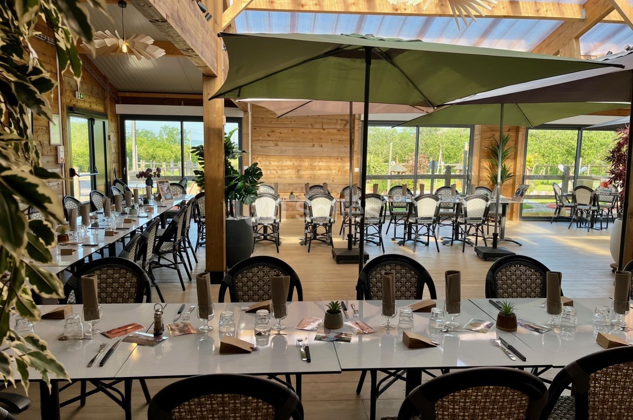 Salle lumineuse avec de grandes baies vitrées, tables dressées et parasols ouverts dans un décor en bois naturel.