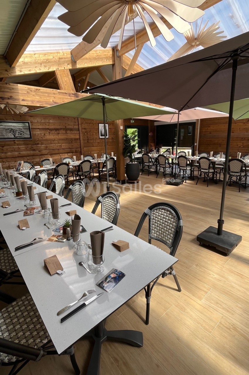 Salle de restaurant lumineuse avec tables dressées, chaises en osier, parasols et décor en bois.