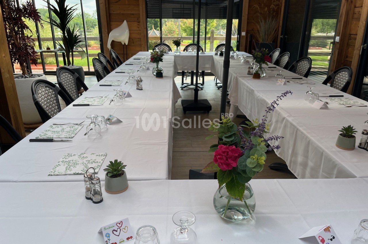 Salle de réunion lumineuse avec tables disposées en U, nappes blanches, décorations florales et vue sur l'extérieur…
