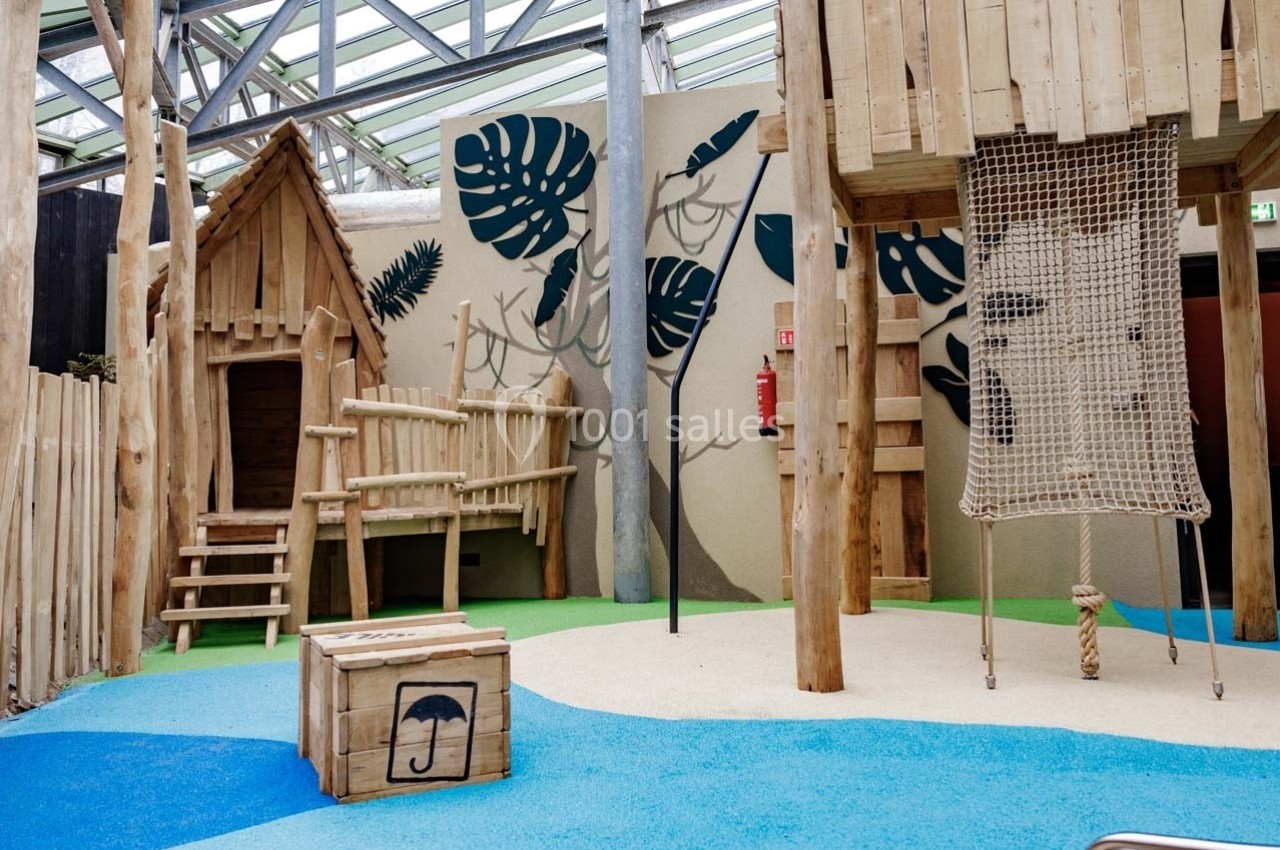 Aire de jeux intérieure en bois avec cabanes, filet d'escalade et décor mural végétal sur sol coloré.