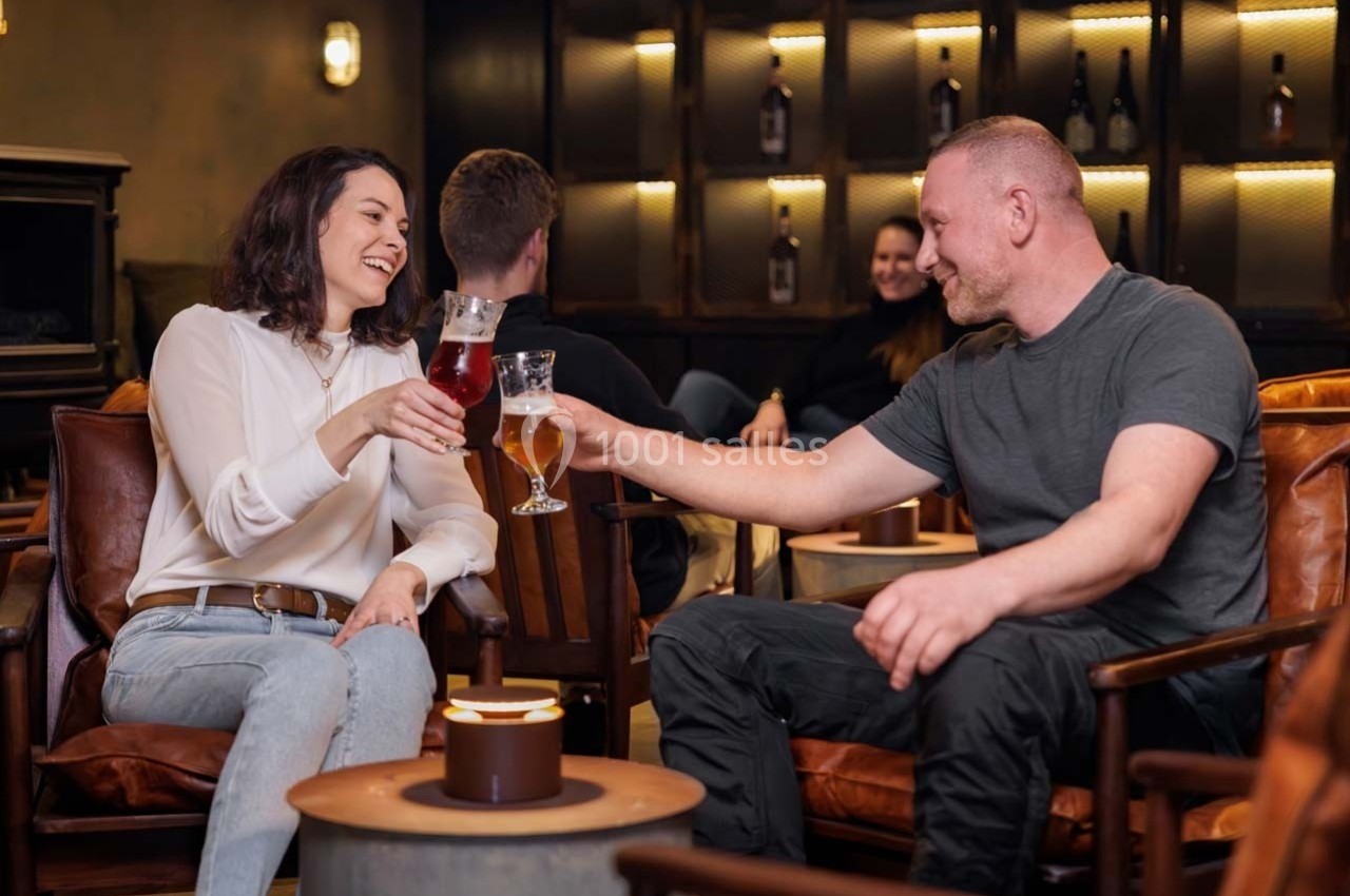 Deux personnes trinquent avec des verres de bière dans un salon chaleureux, entourées d'autres convives.