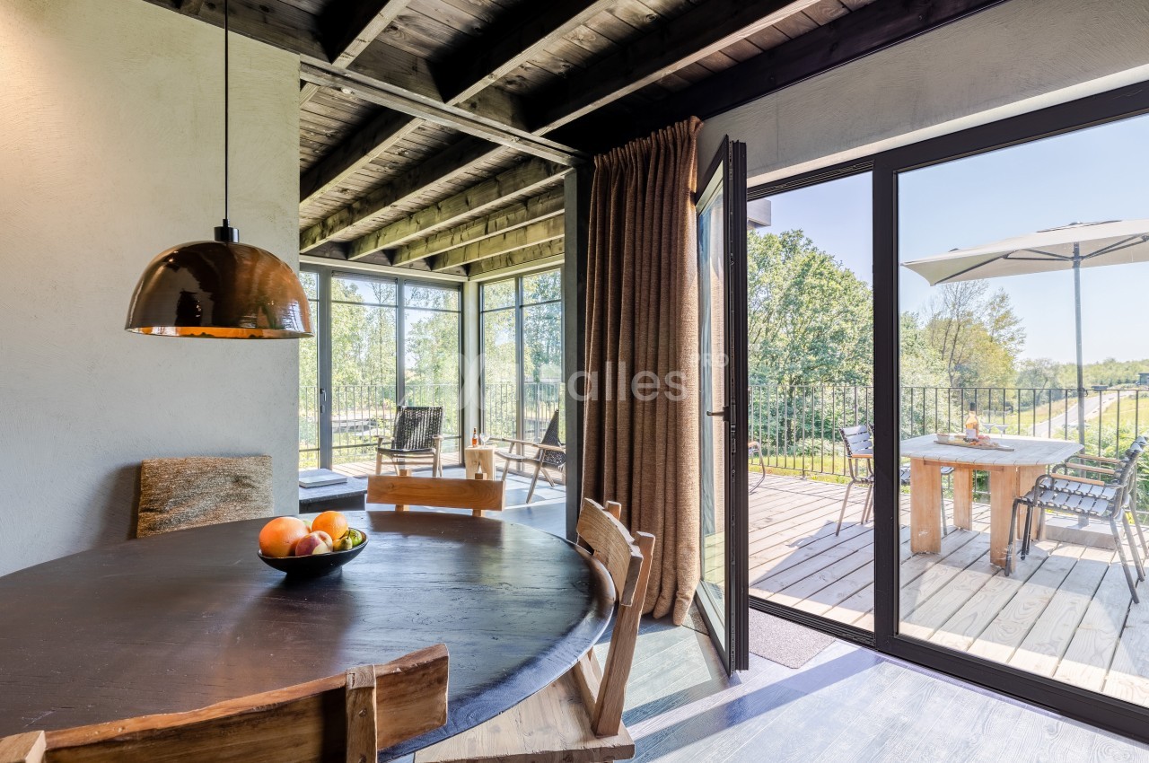 Salle à manger lumineuse avec table en bois, suspension métallique, et accès à une terrasse extérieure avec vue sur la…