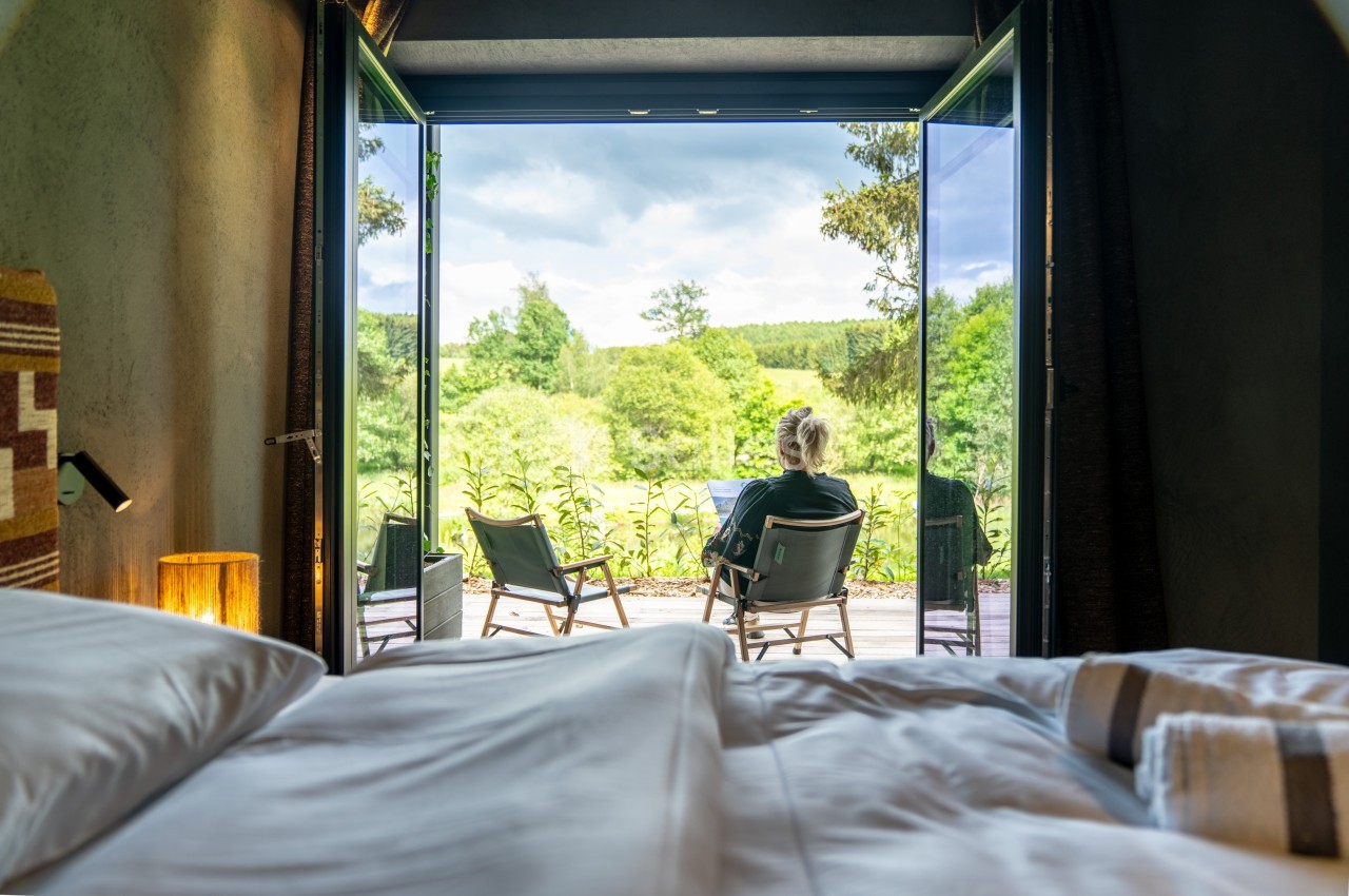 Vue depuis une chambre sur une terrasse avec une personne assise face à un paysage verdoyant sous un ciel nuageux.