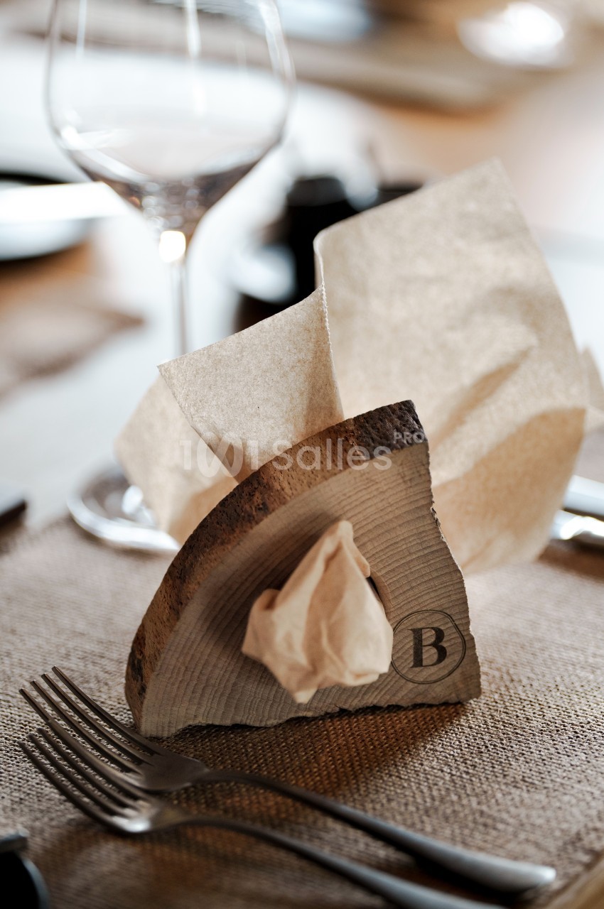 Serviette en papier pliée dans un porte-serviette en bois sur une table dressée avec des couverts et un verre à pied.