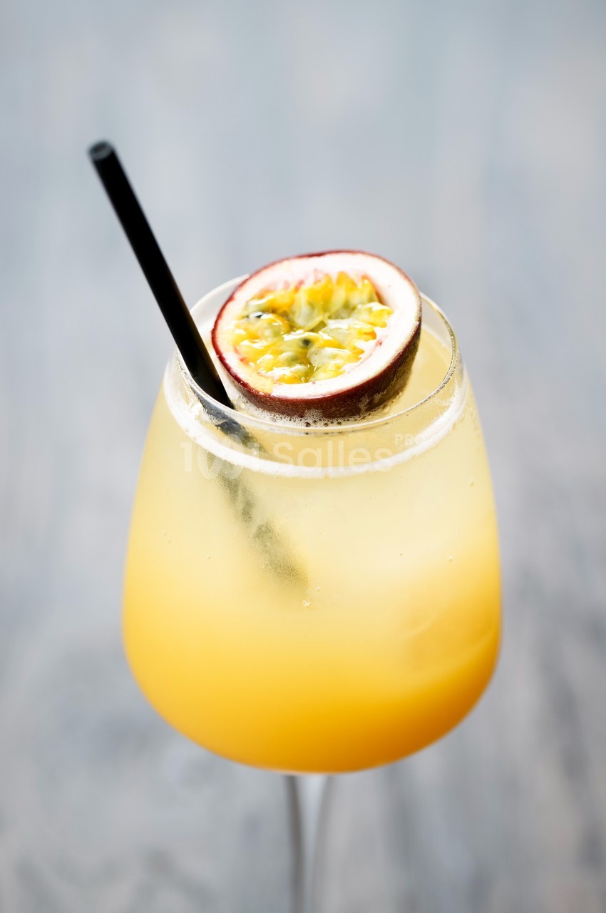 Verre de cocktail jaune garni d'une moitié de fruit de la passion et d'une paille noire, posé sur une surface floue.