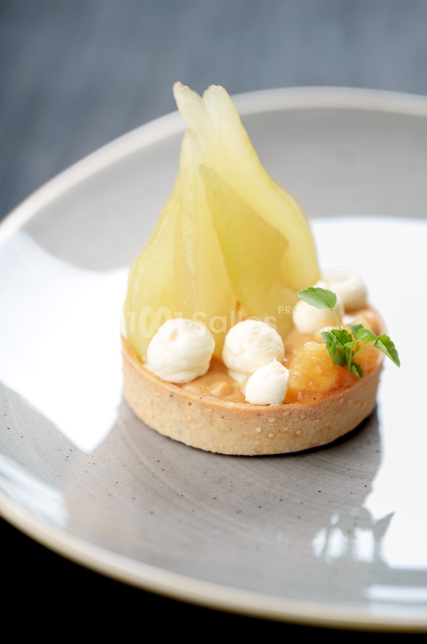 Tartelette garnie de poires pochées, crème légère et décorée de petites feuilles vertes sur une assiette grise.