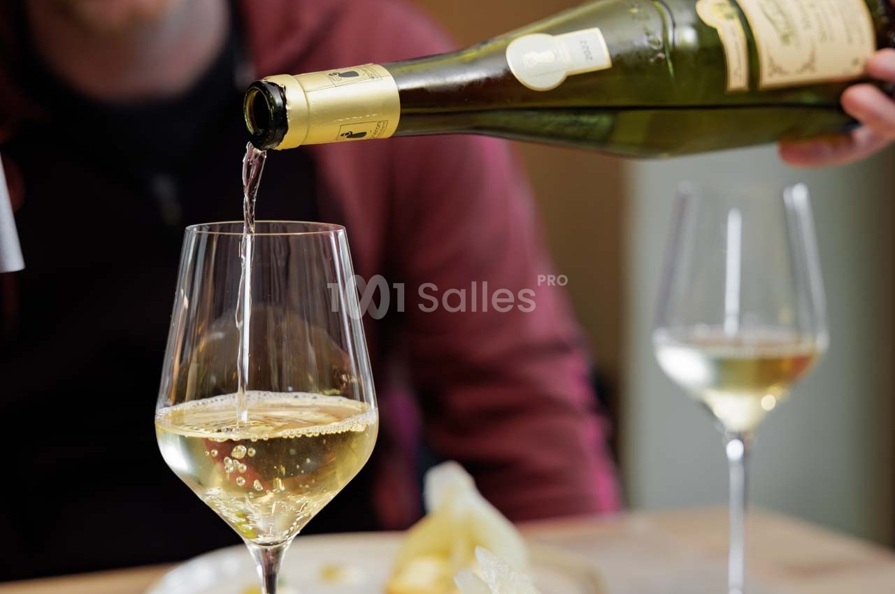 Un verre de vin blanc est servi depuis une bouteille, avec une table et un autre verre en arrière-plan.