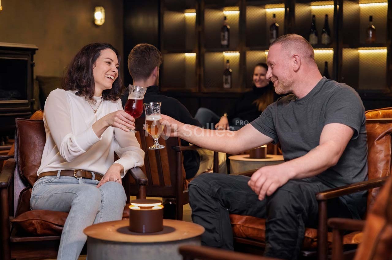 Deux personnes trinquent avec des verres de bière dans un salon chaleureux, entourées d'autres convives.