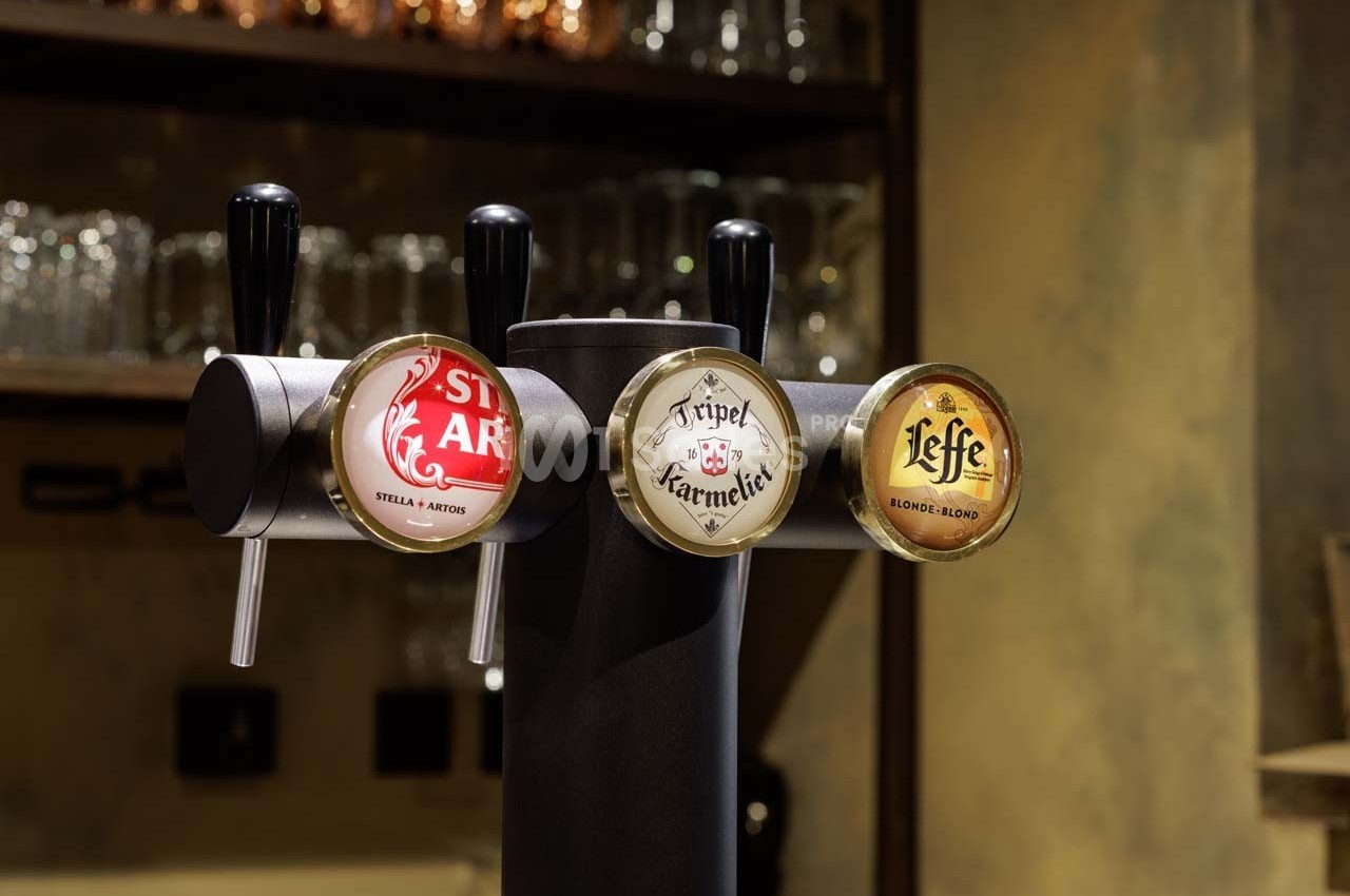 Pompe à bière avec trois tireuses affichant les marques Stella Artois, Tripel Karmeliet et Leffe dans un bar.