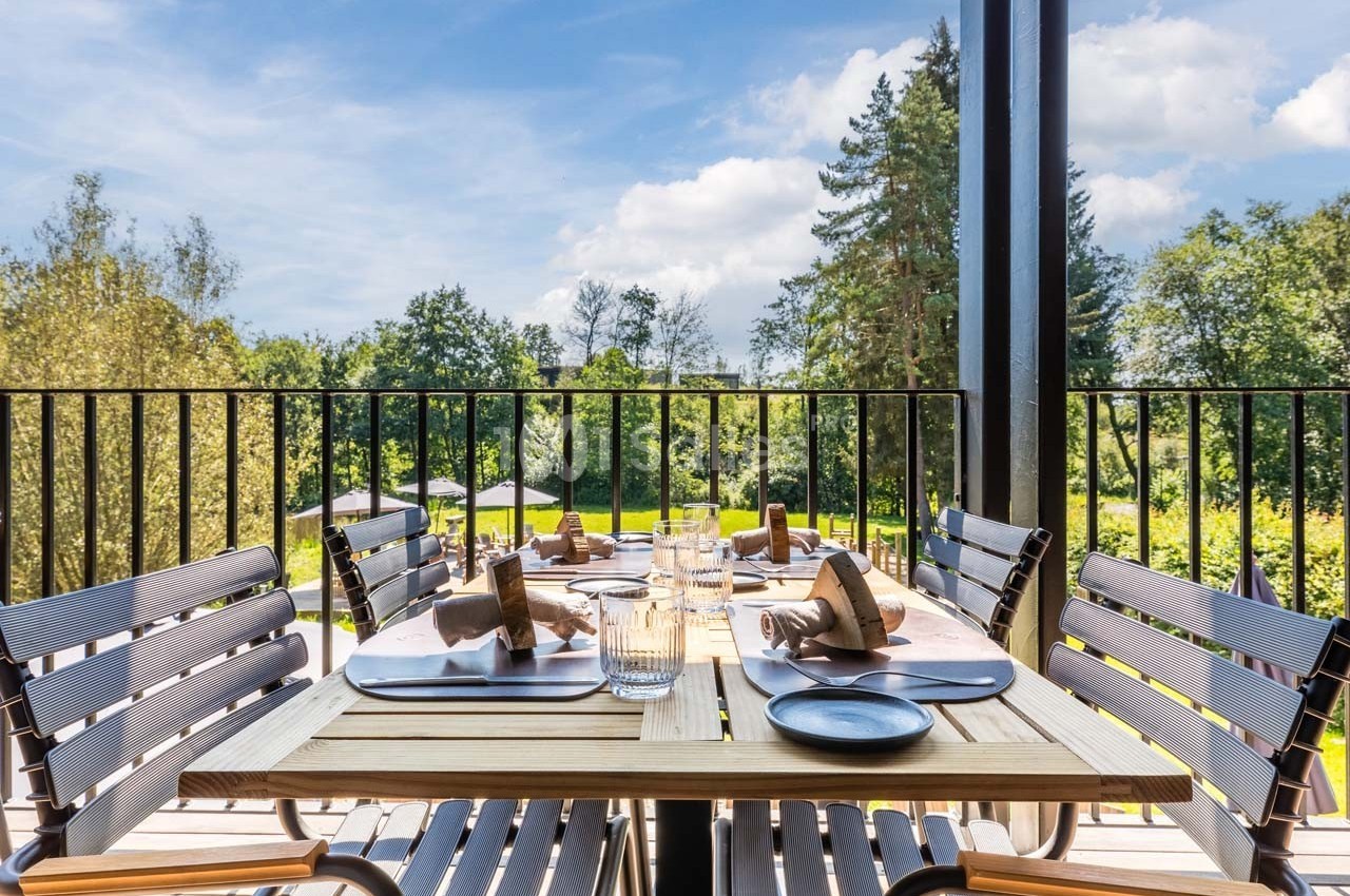 Table en bois dressée avec des assiettes, verres et serviettes sur une terrasse surplombant un jardin verdoyant.