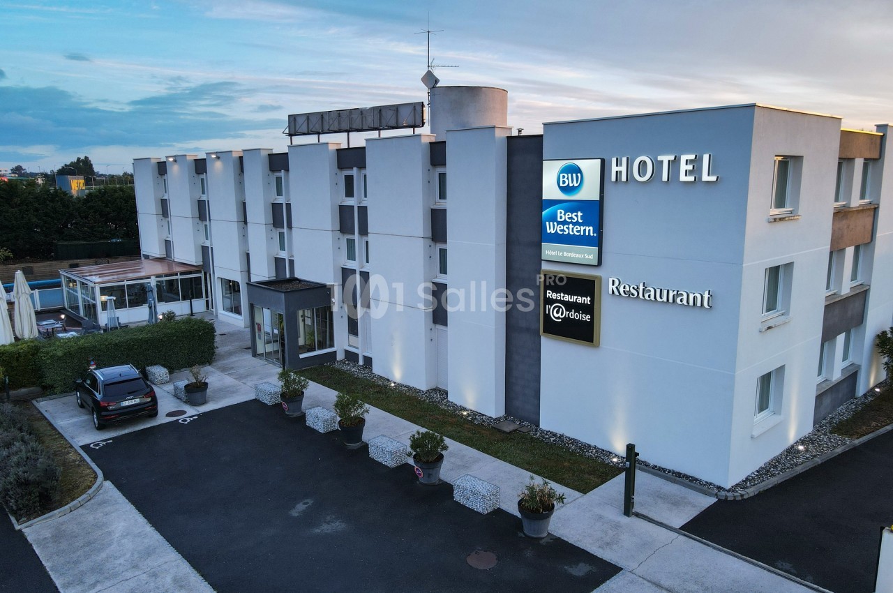 Façade d'un hôtel moderne avec enseignes lumineuses, parking et terrasse extérieure visibles au premier plan.