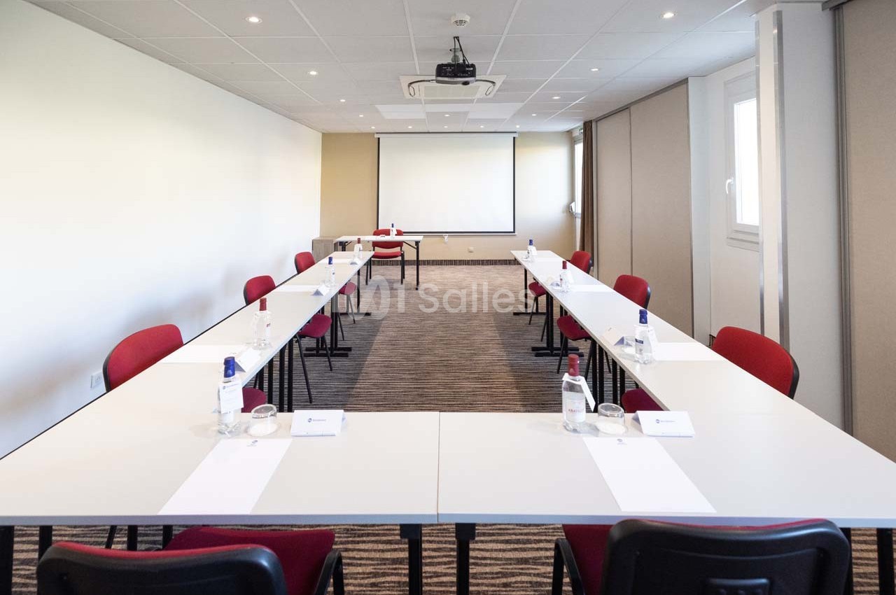 Salle de réunion lumineuse avec tables disposées en U, chaises rouges et écran de projection au fond.