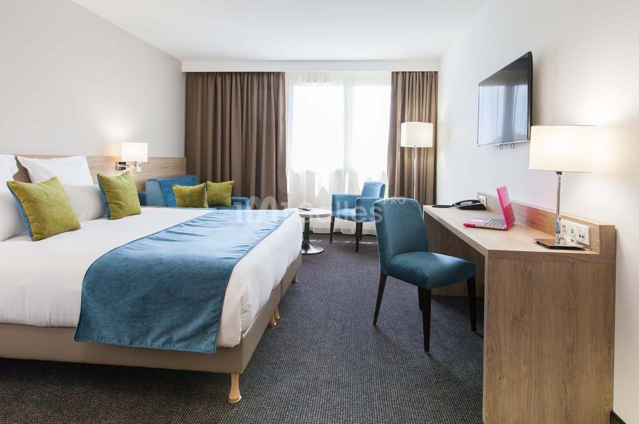 Chambre d'hôtel lumineuse avec lit double, bureau, fauteuils, télévision murale et grandes fenêtres avec rideaux.