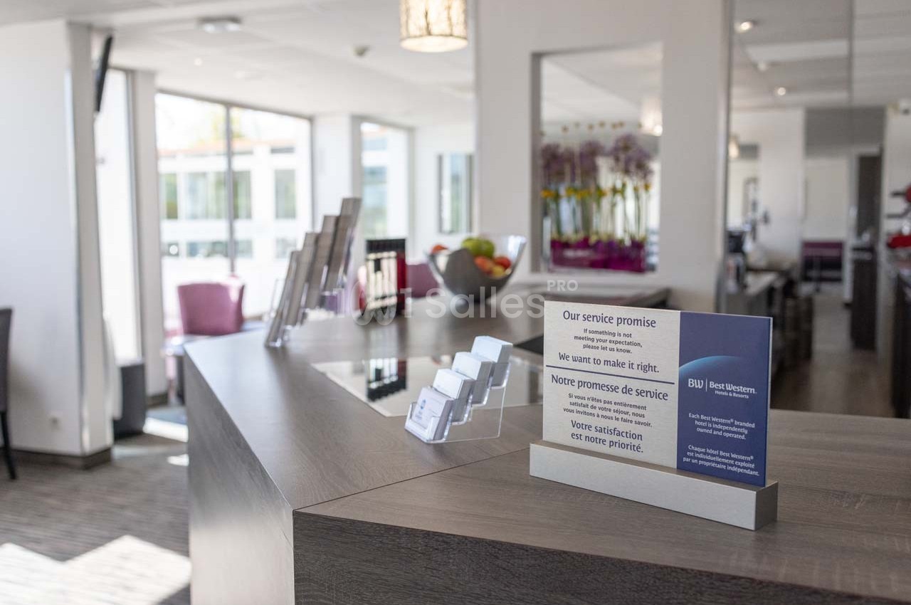 Réception d'hôtel avec un comptoir en bois, présentoirs de cartes de visite et un panneau affichant une promesse de service.