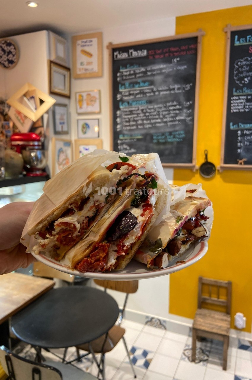 Assiette de sandwichs garnis de légumes grillés et fromage, tenue dans un café au décor chaleureux avec tableau-menu au mur.