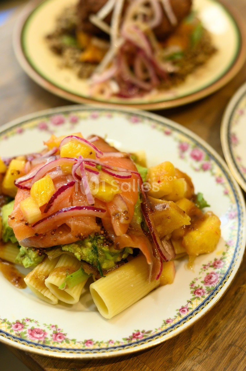 Assiette de pâtes garnie de saumon fumé, mangue, avocat et oignons rouges, servie sur une table en bois.