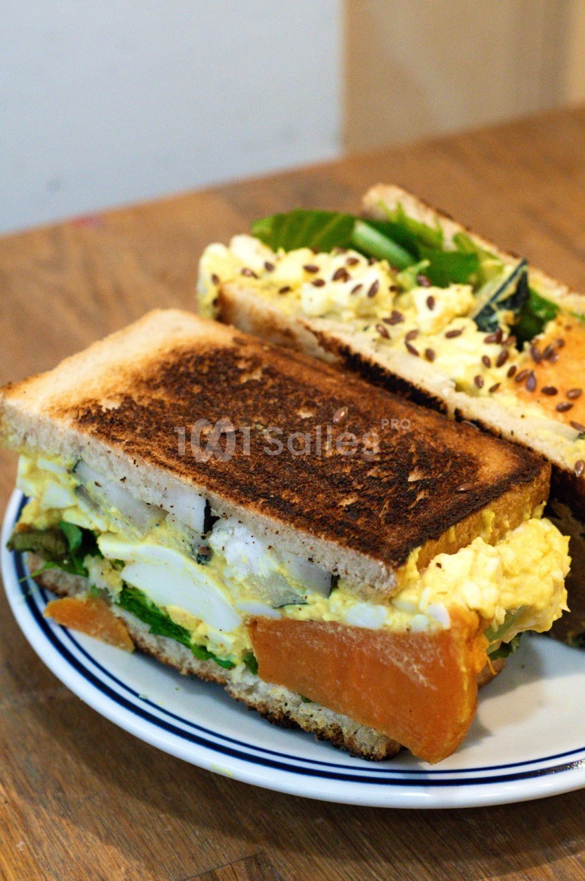 Sandwich garni d'œufs, légumes verts et graines, présenté sur une assiette blanche avec une tranche grillée.
