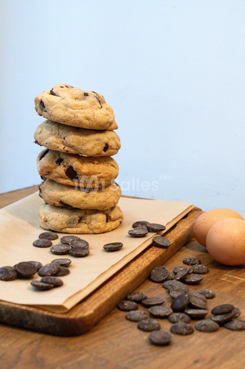 Pile de cookies aux pépites de chocolat sur une planche en bois, entourée d'œufs et de pépites de chocolat.
