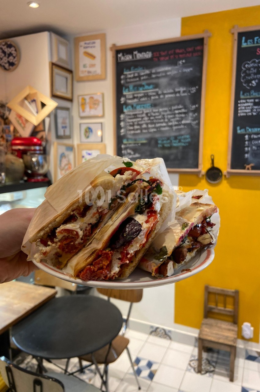 Assiette de sandwichs garnis de légumes grillés et fromage, tenue dans un café au décor chaleureux avec tableau-menu au mur.