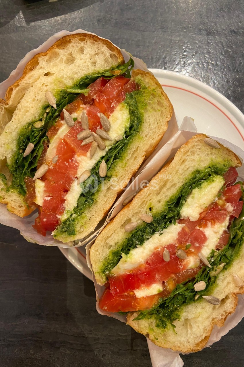 Sandwich garni de tomates, mozzarella, roquette, pesto et graines de tournesol, présenté sur une assiette blanche.