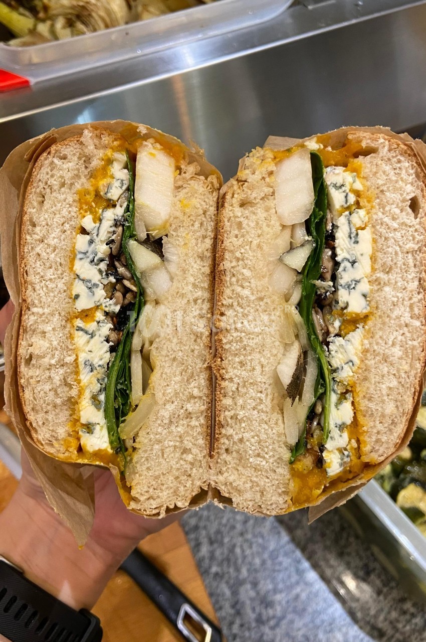 Sandwich coupé en deux, garni de fromage bleu, poires, épinards et autres ingrédients, tenu au-dessus d'un plan de travail.
