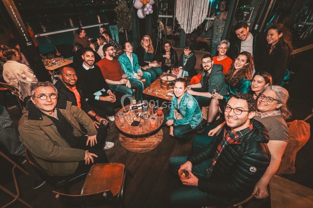 Groupe de personnes assises autour de tables en bois dans un espace convivial, avec des boissons et une ambiance festive.