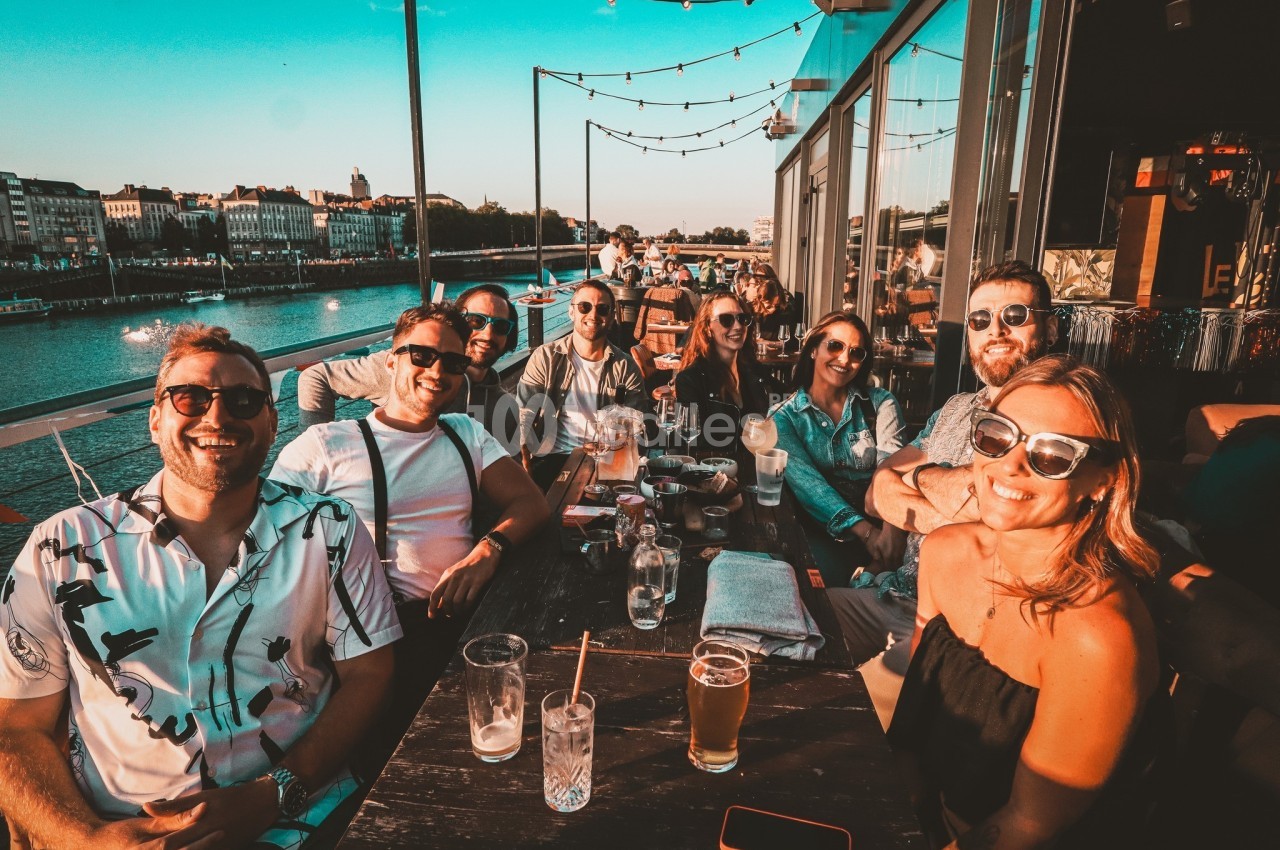 Un groupe de personnes souriantes assises à une table en terrasse au bord d'une rivière, par temps ensoleillé.