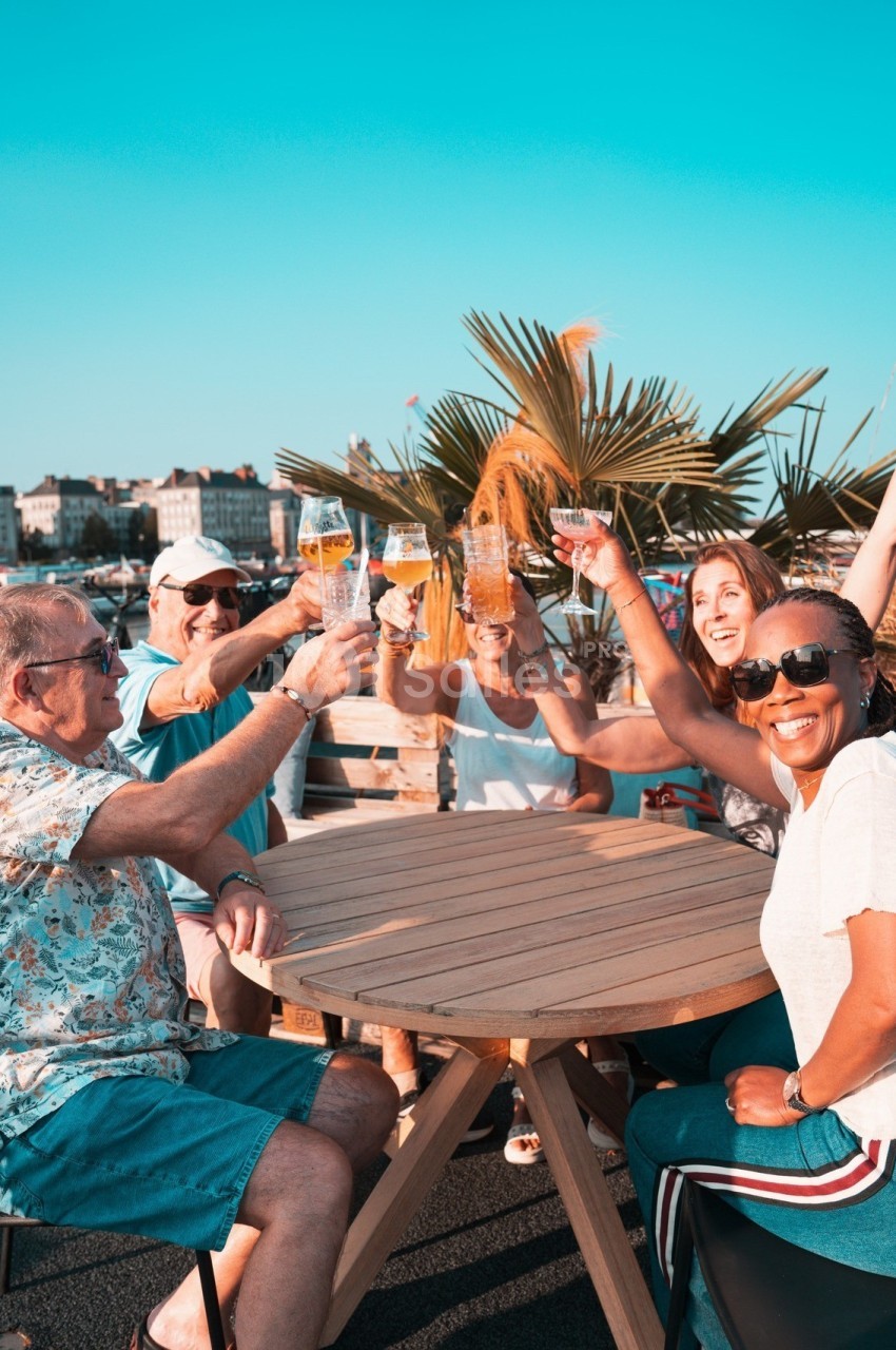 Des personnes souriantes trinquent avec des verres autour d'une table en extérieur, sous un ciel bleu.