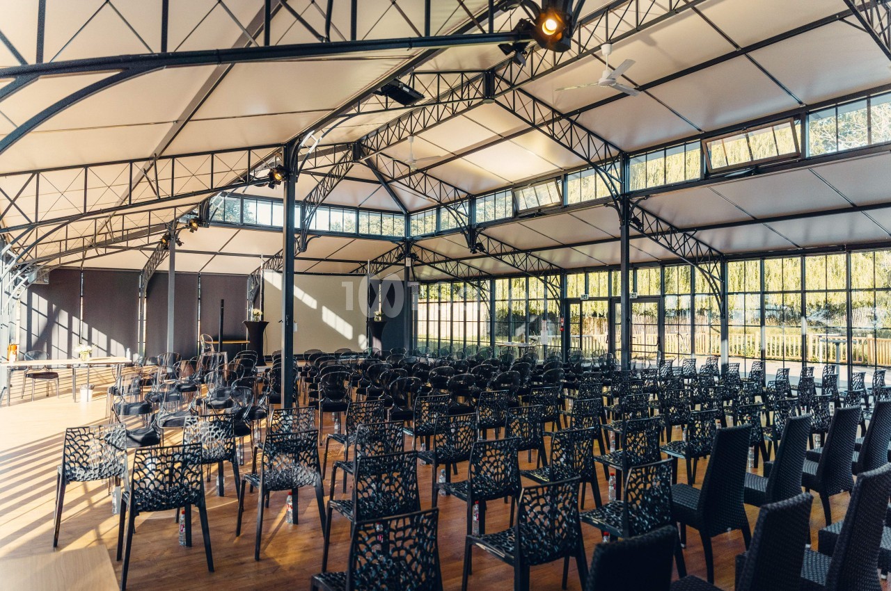 Salle lumineuse avec toit en verrière, rangées de chaises noires disposées face à une scène en bois.