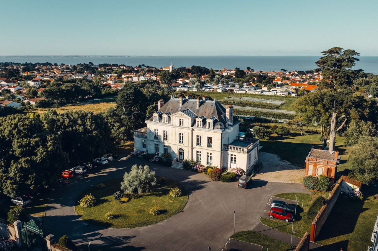 Vue aérienne d'un manoir entouré de jardins, avec une petite ville et la mer en arrière-plan.