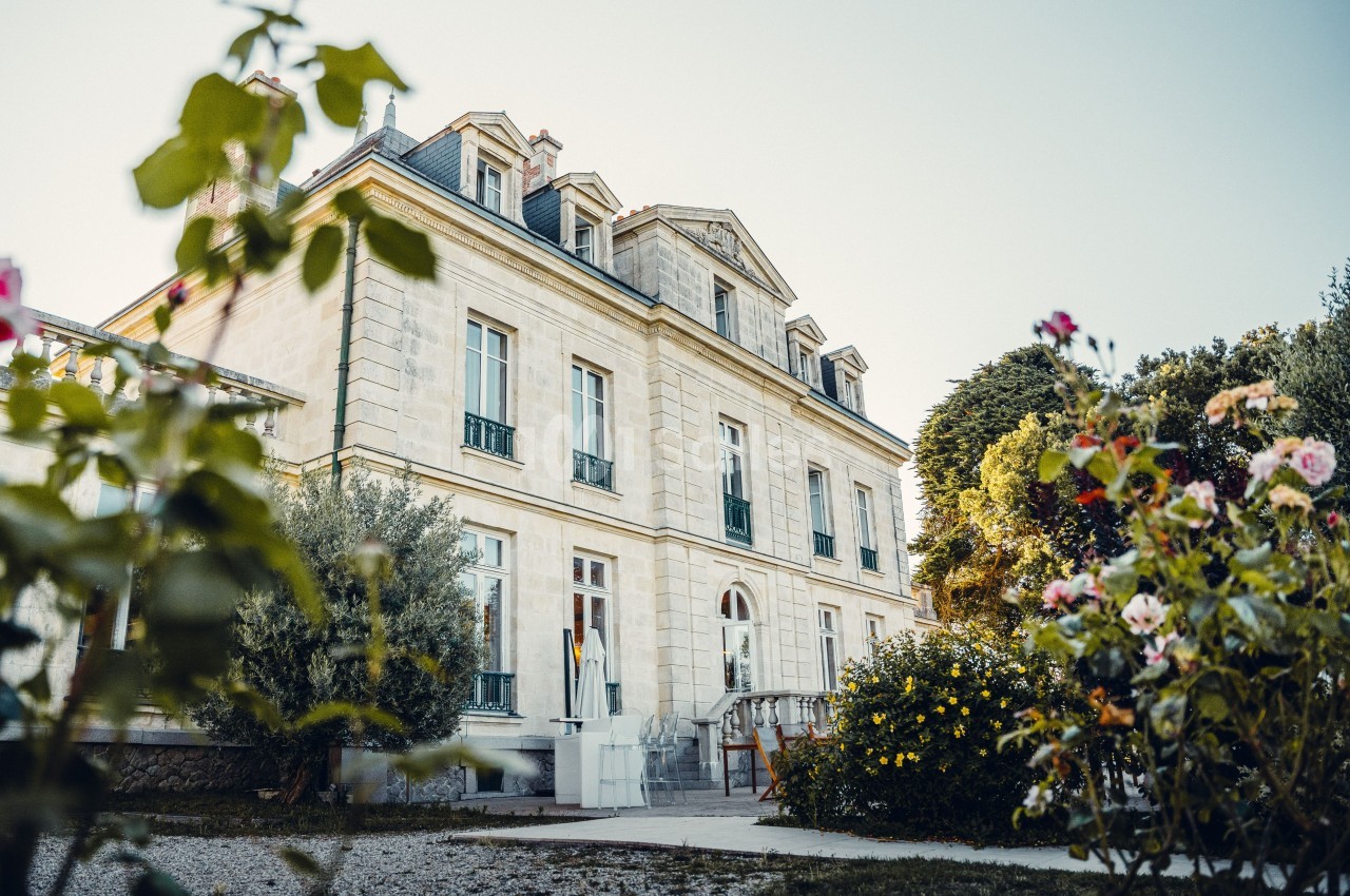 Façade d'un manoir en pierre entouré de fleurs et de verdure, sous un ciel dégagé.