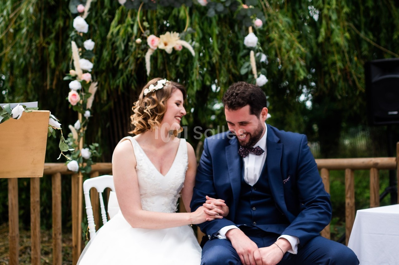 Un couple assis, souriant et se tenant la main lors d'une cérémonie de mariage en extérieur.