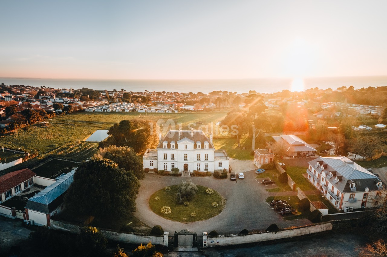 Vue aérienne d'un manoir entouré de jardins, avec un village et la mer en arrière-plan au coucher du soleil.