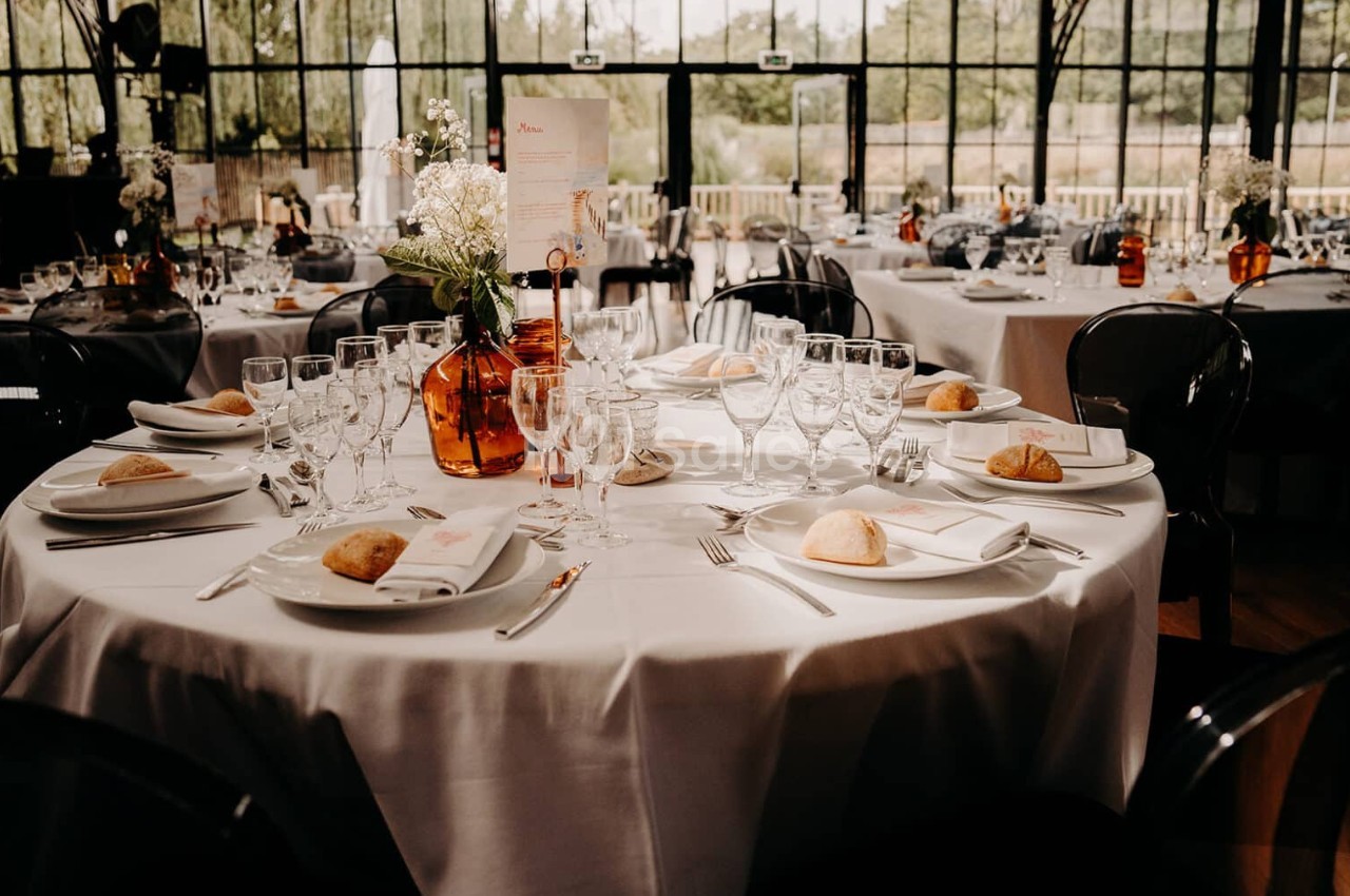 Tables de réception élégamment dressées avec nappes blanches, vaisselle, verres et décorations florales.