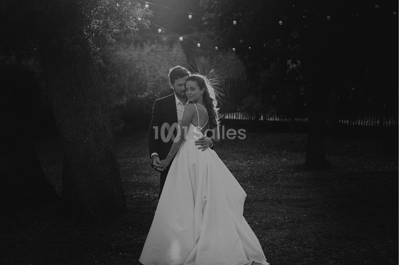 Un couple en tenue de mariage pose dans un jardin sombre, éclairé par des guirlandes lumineuses suspendues.