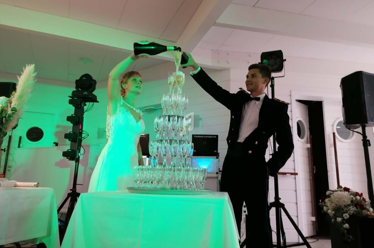 Un couple en tenue de cérémonie verse du champagne dans une pyramide de verres lors d'une réception festive.