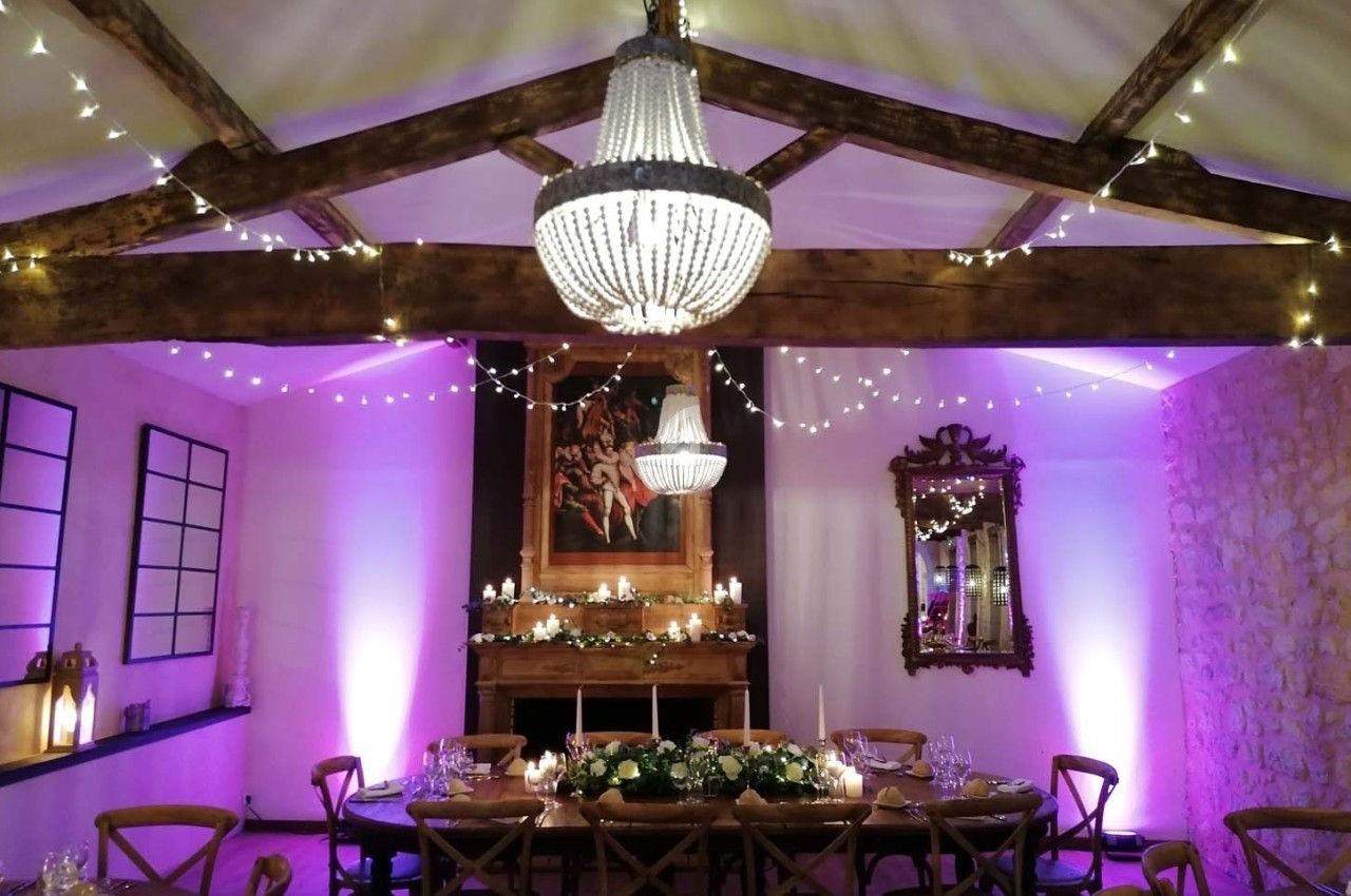 Salle décorée pour un dîner, avec table ornée de fleurs, guirlandes lumineuses et lustre en cristal suspendu.