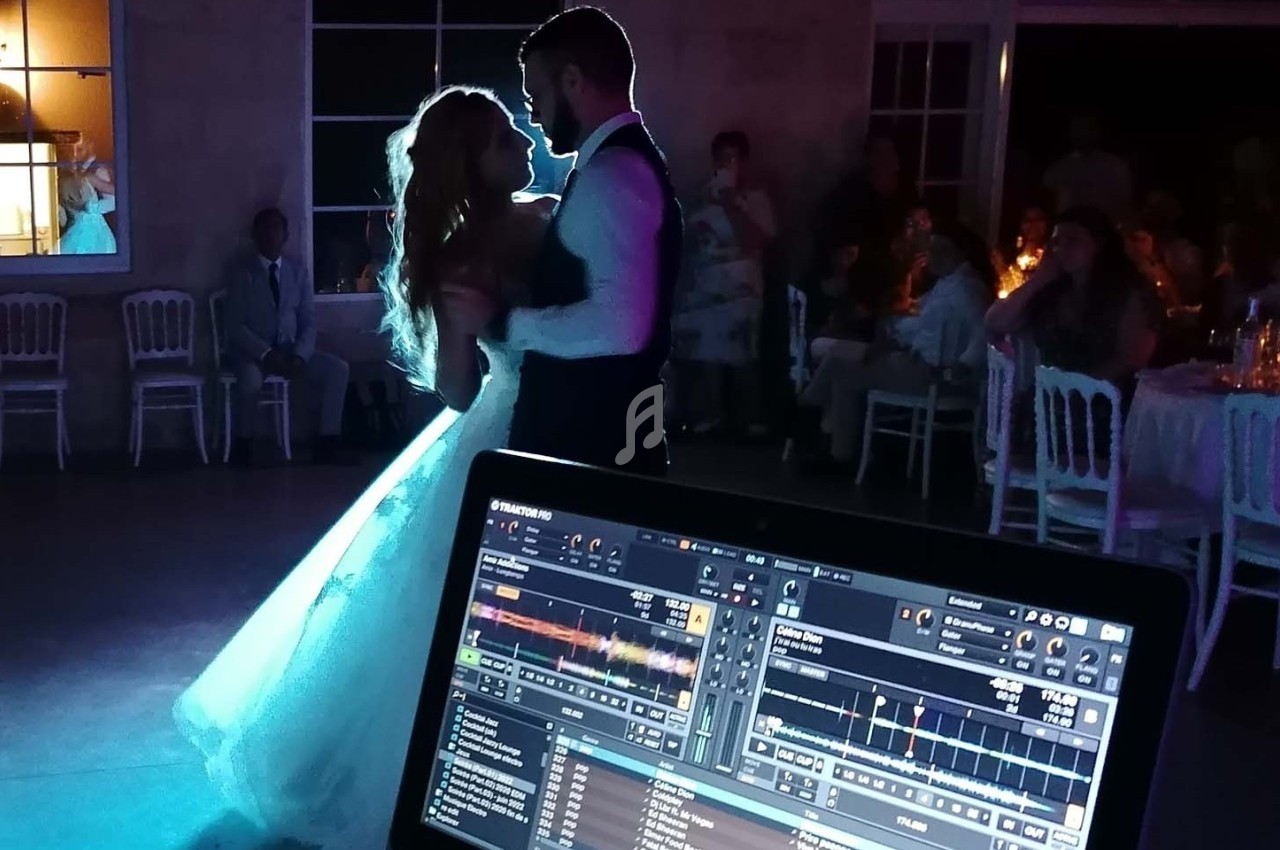Un couple danse lors d'un mariage, éclairé par des lumières tamisées, avec une table de mixage visible au premier plan.