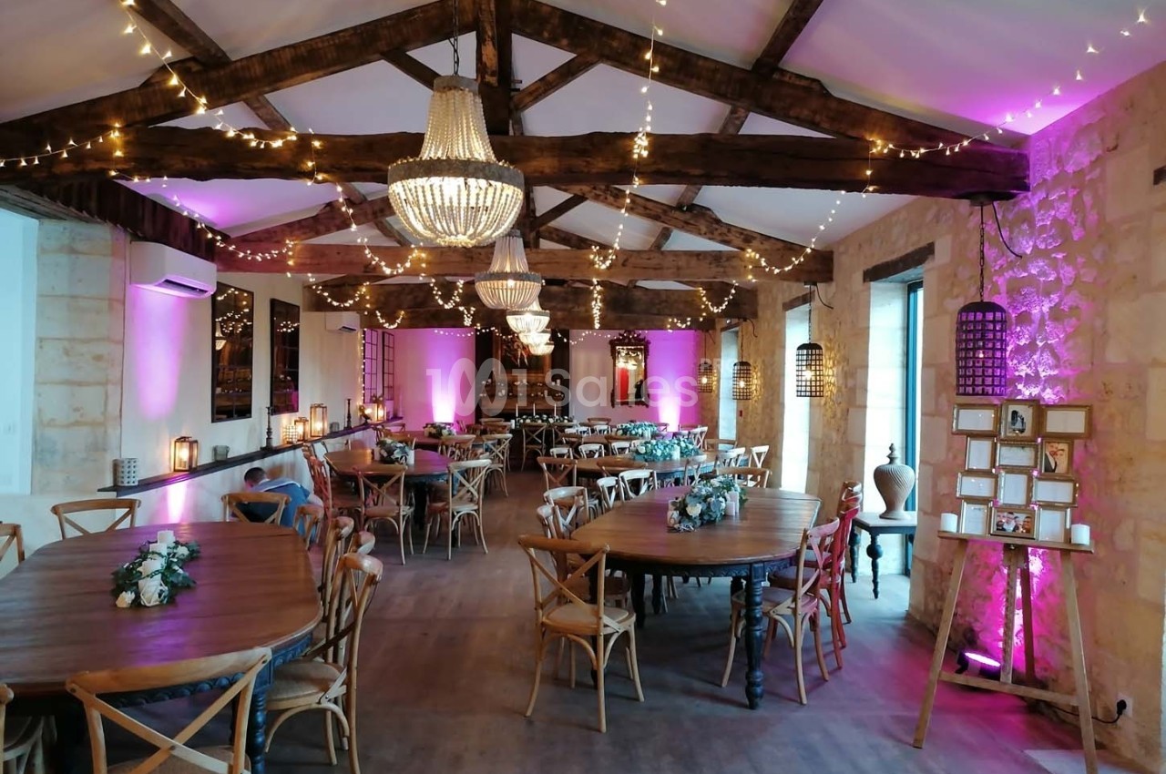 Salle de réception avec poutres apparentes, tables en bois décorées de fleurs et éclairages roses et chaleureux.