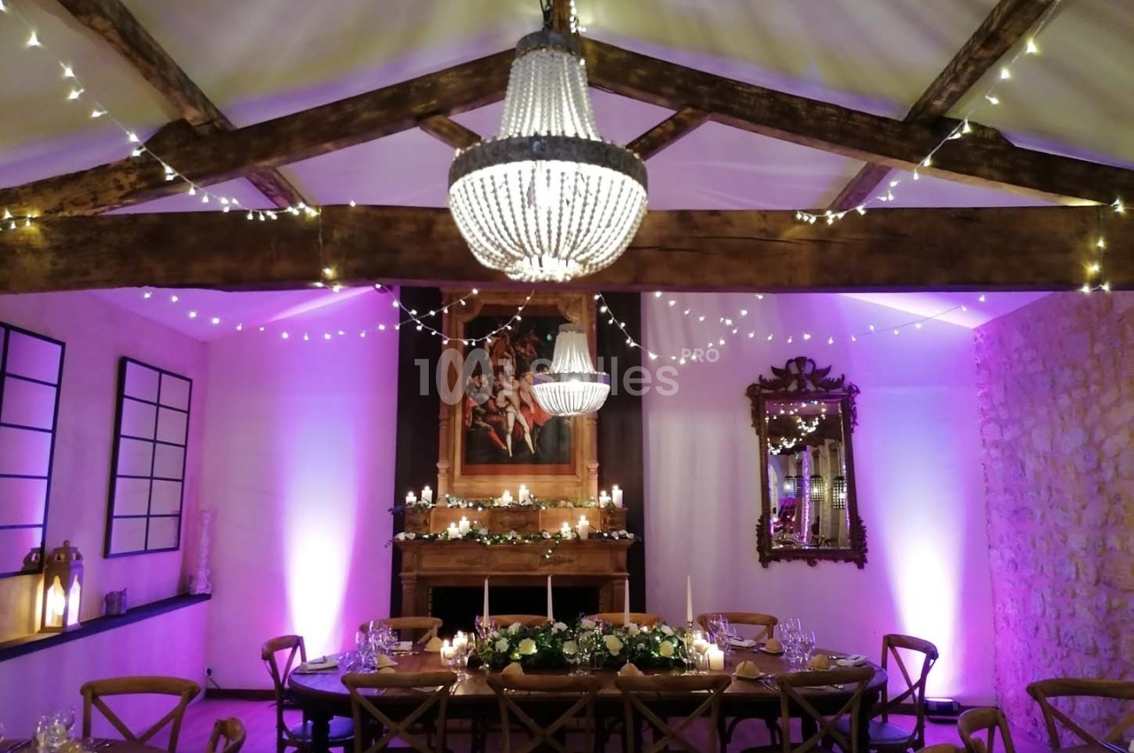 Salle décorée pour un dîner, avec table dressée, guirlandes lumineuses, lustres et éclairage d'ambiance violet.