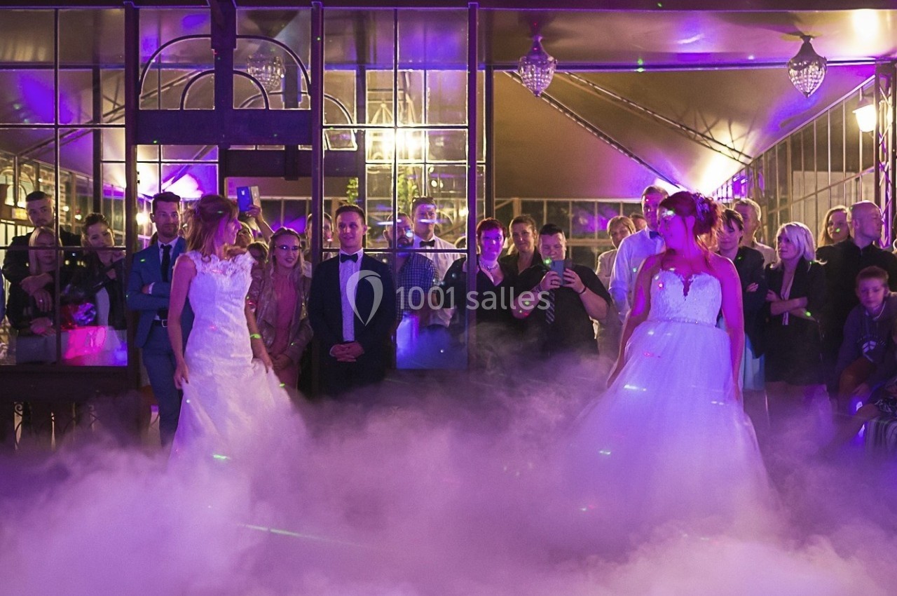 Deux mariées en robe blanche dansent au milieu d'une piste entourée de fumée et de convives sous des lumières colorées.
