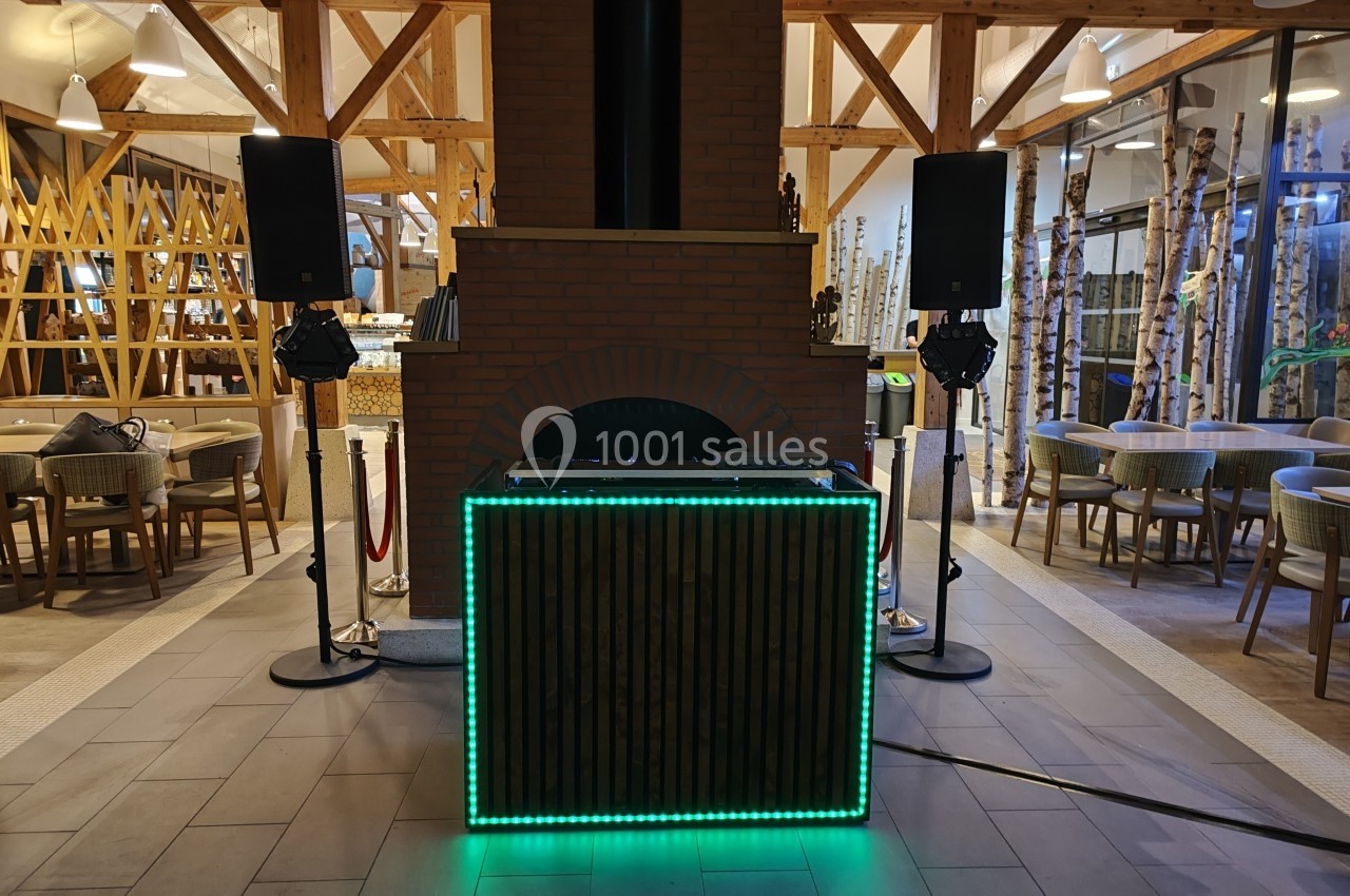 Table de DJ éclairée par des LED vertes, entourée de haut-parleurs, dans un espace intérieur avec structure en bois.