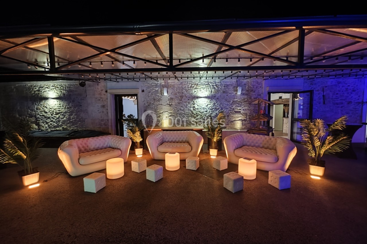 Espace extérieur aménagé avec canapés, tables basses lumineuses, plantes et éclairage chaleureux devant un mur en pierre.