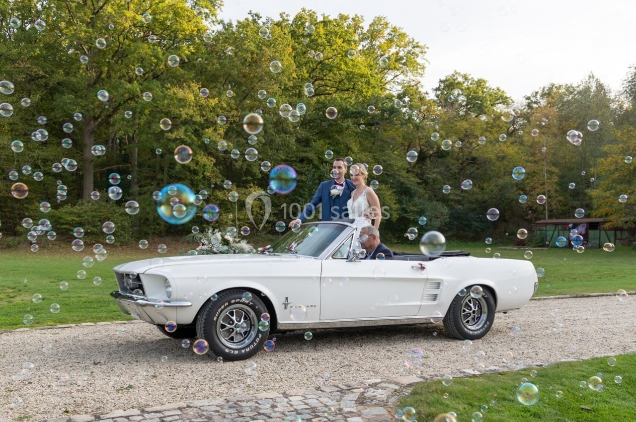 Un couple en tenue de mariage pose dans une voiture décapotable blanche entourée de bulles, en extérieur verdoyant.